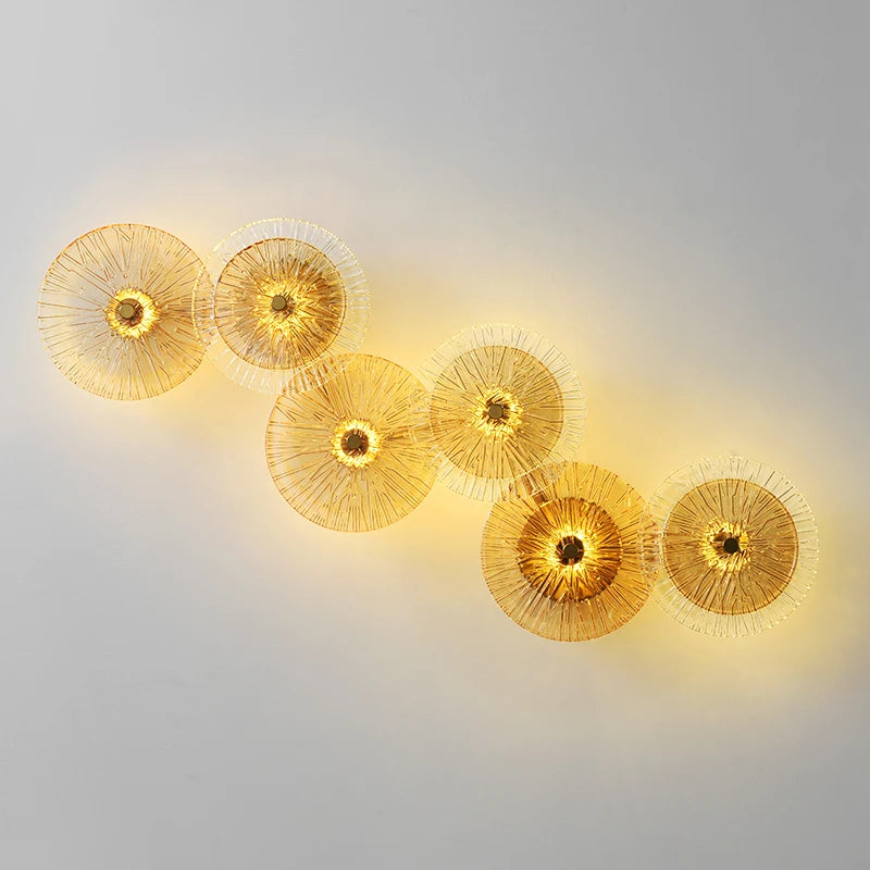 Six Amber Deco Wall Sconces, featuring circular gold wire designs, are mounted in a staggered line on a plain gray wall, casting a warm yellow glow.