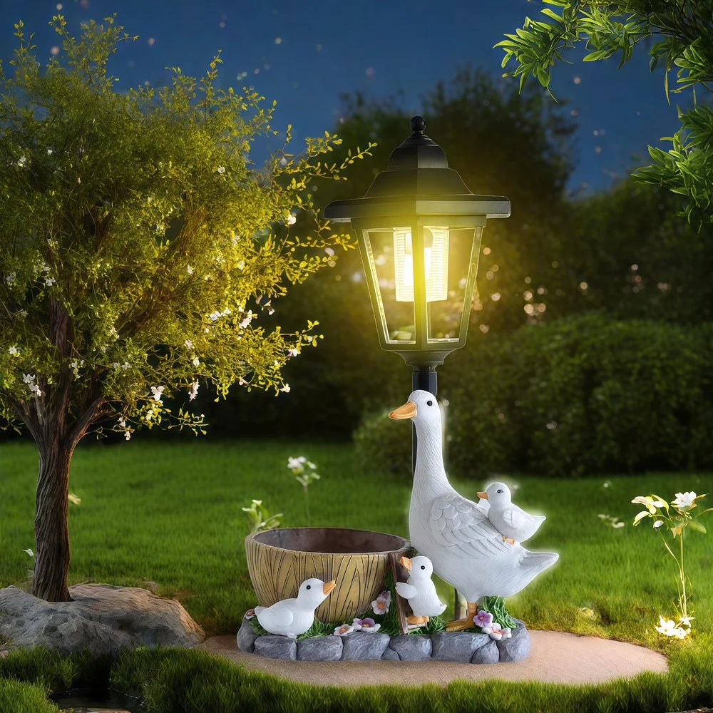 The Solar Goose Lamp, illuminated at night, features a charming goose with three goslings resting beside a tree and a small wooden barrel on a grassy lawn, bringing whimsical charm to your cottage garden decor.
