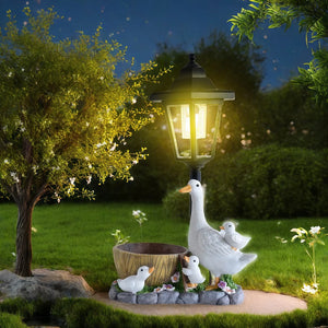 The Solar Goose Lamp, illuminated at night, features a charming goose with three goslings resting beside a tree and a small wooden barrel on a grassy lawn, bringing whimsical charm to your cottage garden decor.
