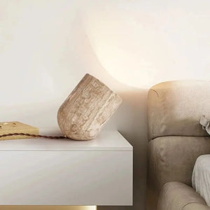 A Natural Stone Lamp, sculptural in form, adds an artistic touch to the serene setting on a white table beside a beige sofa, accompanied by a beige ceramic vase lying on its side and a wooden item with a cord.