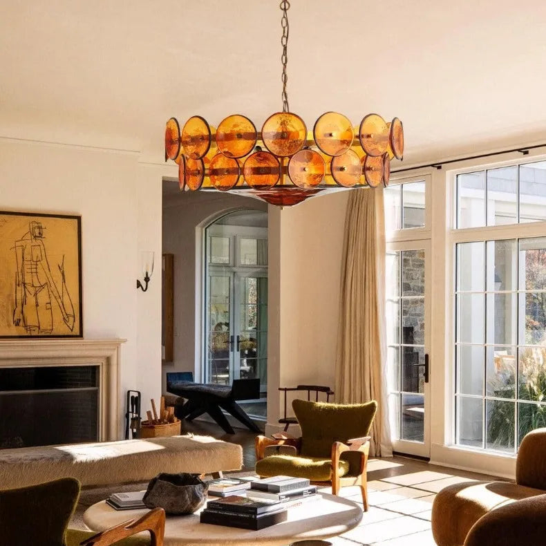 A sunlit living room with modern furniture, a round coffee table, olive green chairs, a stone fireplace, wall art, and a striking Italian Retro Pendant Light. Large windows bathe the space in natural light.