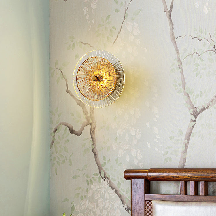 The Amber Deco Wall Sconce, with its circular shape, golden glow, and delicate glass disc design, is mounted on a wallpapered wall with a tree branch pattern above a wooden headboard.