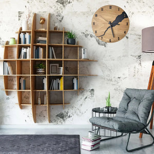 A modern living room showcasing a wood bookshelf with a natural wood finish, a gray cushioned chair, and a side table adorned with books. The Wooden Round Wall Clock introduces charm to the space while textured walls highlight the room's unique crack design. A tall lamp rounds out the cozy ambiance.