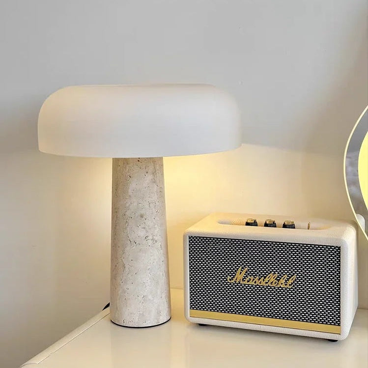 A Mushroom Desk Lamp with a white shade and a natural stone base provides organic lighting to your desk, placed beside a cream-colored Marshall speaker on a white surface.