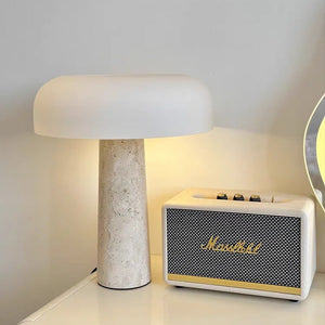 A Mushroom Desk Lamp with a white shade and a natural stone base provides organic lighting to your desk, placed beside a cream-colored Marshall speaker on a white surface.