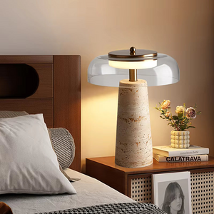 The Mushroom Desk Lamp, featuring a natural stone base and glass dome, is placed on a wooden nightstand beside the bed, alongside a vase of flowers, two books, and a black-and-white photo—adding an inviting touch of organic desk lighting to the cozy setting.
