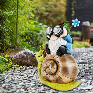 A whimsical Solar Gnome Snail Light featuring a garden gnome with binocular-like eyes and a blue hat perched atop a large snail brightens your stone path, surrounded by lush greenery. This charming piece of outdoor decor enhances your landscape both day and night with its delightful design and gentle solar-powered glow.