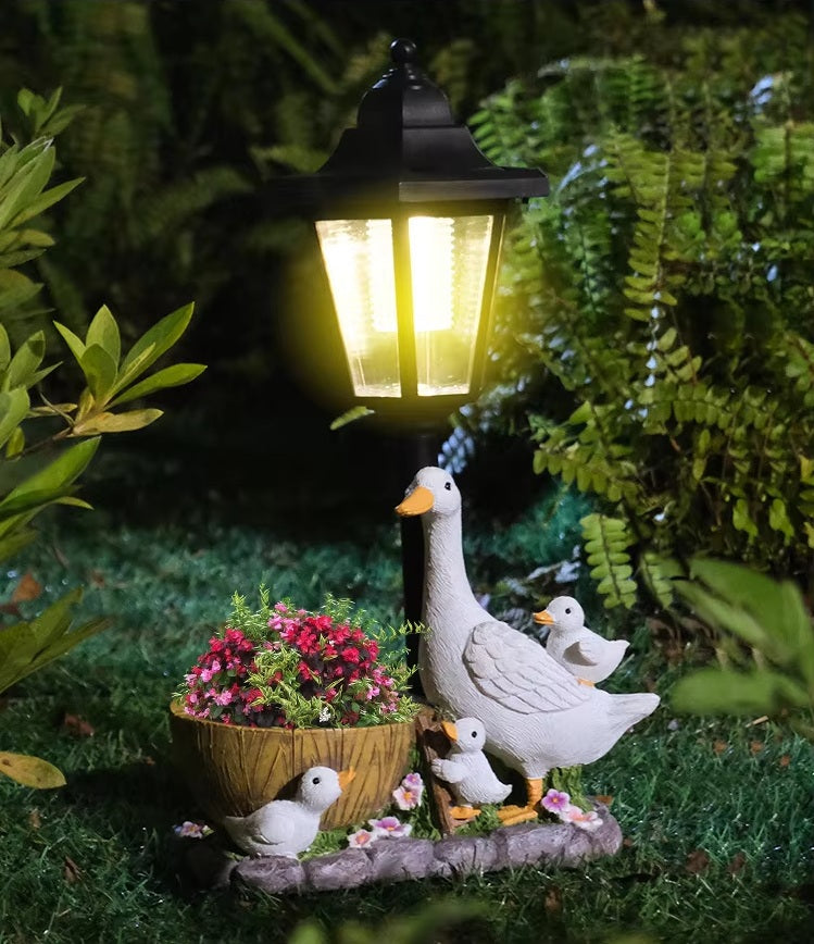 The Solar Goose Lamp brightens your garden with its delightful design, featuring a goose and three gosling statues beside a pot of pink flowers and lush green plants—making it the perfect decor for any cottage-style garden.