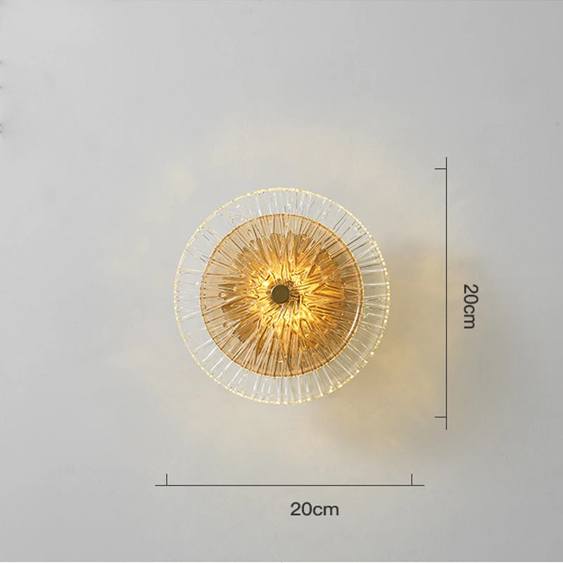 The Amber Deco Wall Sconce, with a gold center and clear glass disc, measures 20 x 20 cm and is stylishly displayed on a plain light-colored wall.