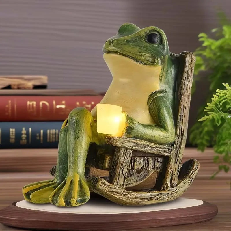 A Solar-powered Rocking Chair Frog Light features a frog figurine seated in a wooden rocking chair, holding a glowing yellow cube, with books and a plant in the background.