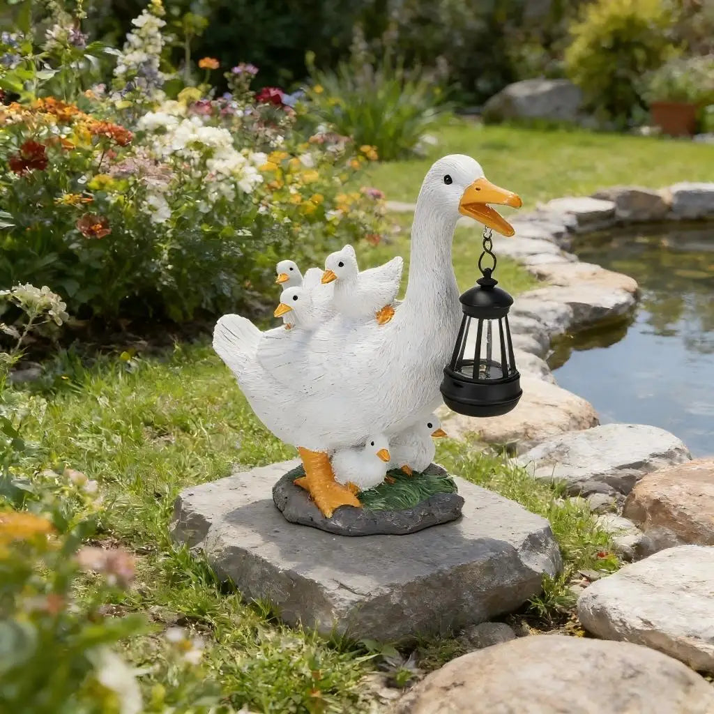 The Solar Duck Lamp, featuring a white duck statue with three ducklings and a hanging black solar-powered lantern, stands on a stone by a pond amidst colorful flowers and lush garden greenery.