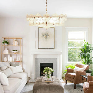 A bright living room with a white sofa, two brown leather chairs, a tufted ottoman, fireplace, artwork, a gold shelf, plants, and an elegant Italian Retro Pendant Light as the stunning centerpiece.