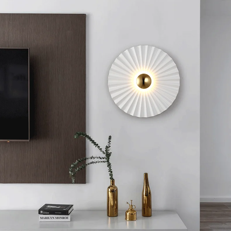 A modern living room wall featuring the Origami Circle Wall Light, part of a TV screen, and a white shelf with gold decorative items and a plant.