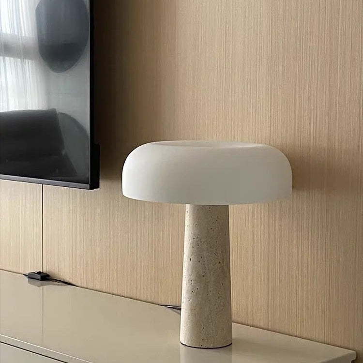 The Mushroom Desk Lamp, featuring a natural stone base and a white dome-shaped shade, sits on a beige surface beside a wall-mounted flat-screen TV, providing elegant and organic lighting ideal for any contemporary space.