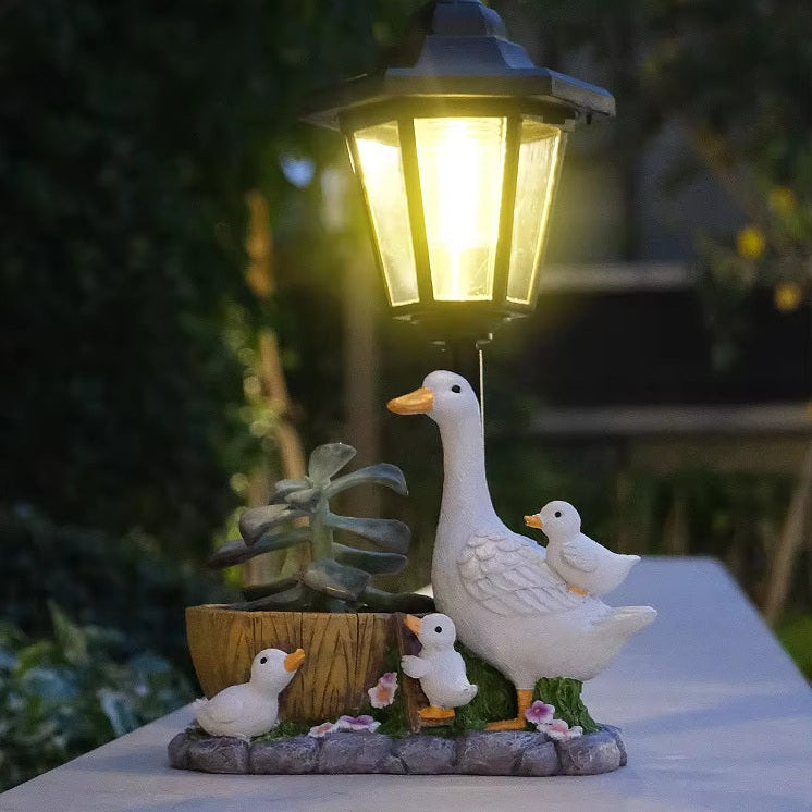 Brighten your cottage garden with the Solar Goose Lamp, a delightful outdoor light featuring a charming white goose, three ducklings, a small plant, and a wooden barrel at its decorative base. This whimsical lamp creates a peaceful atmosphere as it illuminates your garden at dusk.