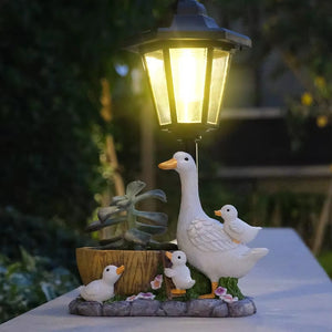 Brighten your cottage garden with the Solar Goose Lamp, a delightful outdoor light featuring a charming white goose, three ducklings, a small plant, and a wooden barrel at its decorative base. This whimsical lamp creates a peaceful atmosphere as it illuminates your garden at dusk.