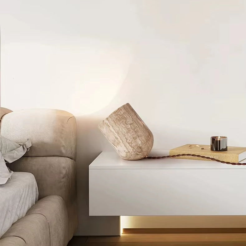 A modern bedroom features a beige upholstered bed, a white floating nightstand, a tilted stone vase, a wooden tray, a candle, and a beaded string against a plain white wall. Accenting the space is the Natural Stone Lamp, crafted from yellow cave stone, which adds an artistic touch and subtle warmth to the serene setting.