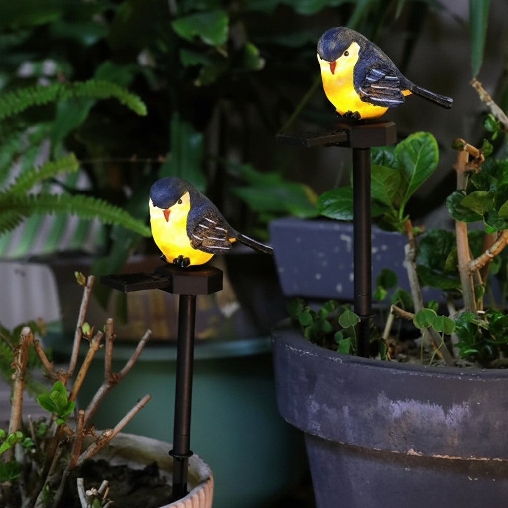 Two Solar Bird Lights in yellow and black are mounted on stakes in potted plants, surrounded by green foliage.