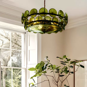 An Italian Retro Pendant Light with green glass discs hangs from the ceiling in a bright room featuring cream walls, a large window, and a leafy indoor plant below.
