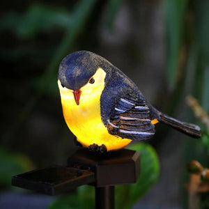 A realistic yellow and blue Solar Bird Light, perfect as songbird decor, is perched on a black stand with greenery blurred in the background.
