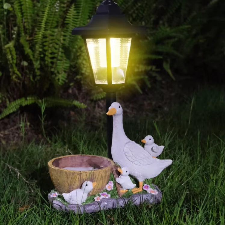 The Solar Goose Lamp illuminates a charming goose planter with three goslings nestled on grass, creating delightful garden decor ideal for a cottage garden setting, beautifully highlighted by ferns in the background at night.