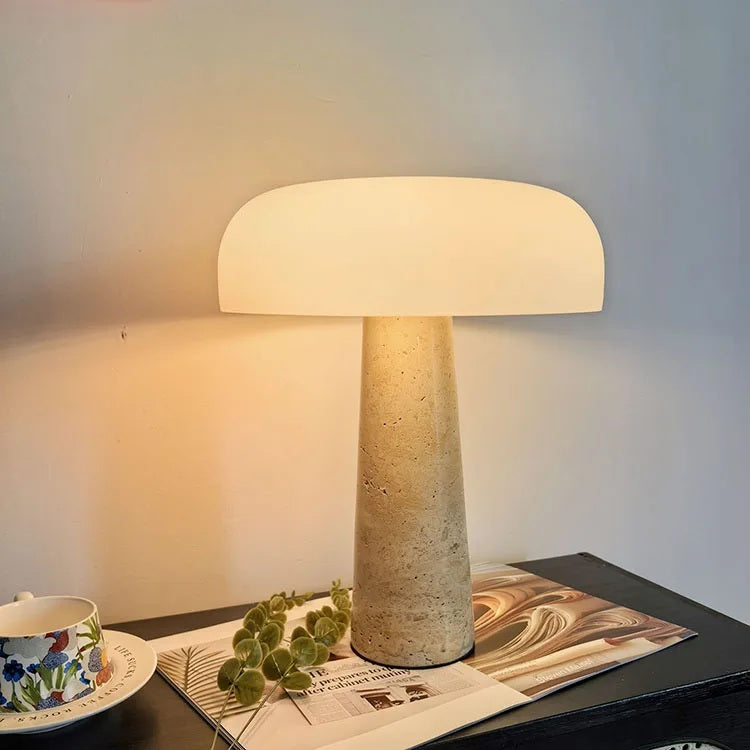 The Mushroom Desk Lamp, featuring a natural stone base and a white shade, is illuminated on a dark table beside an open magazine, a floral-patterned cup, and a sprig of greenery, adding organic lighting to the cozy scene.
