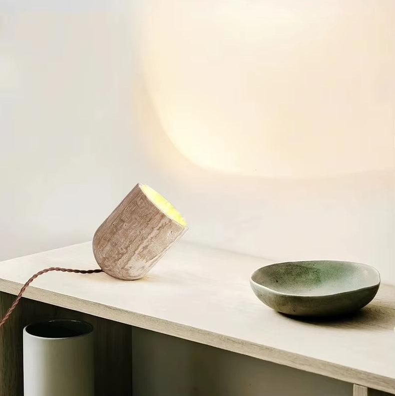 A sculptural Natural Stone Lamp, crafted from yellow cave stone in a small cylindrical shape, casts a warm glow on a wooden surface beside a round greenish-grey ceramic dish. Its graceful cord extends from the base.