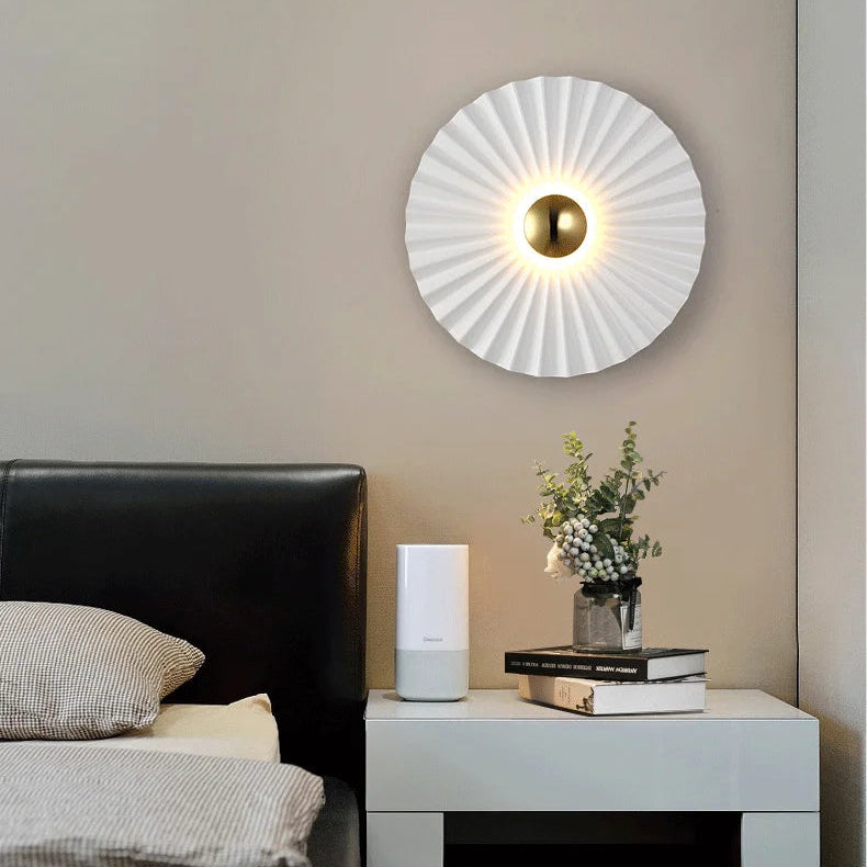 A modern bedroom features the Origami Circle Wall Light above a nightstand with books, a potted plant, and an electronic device, paired with a black headboard and striped bedding.