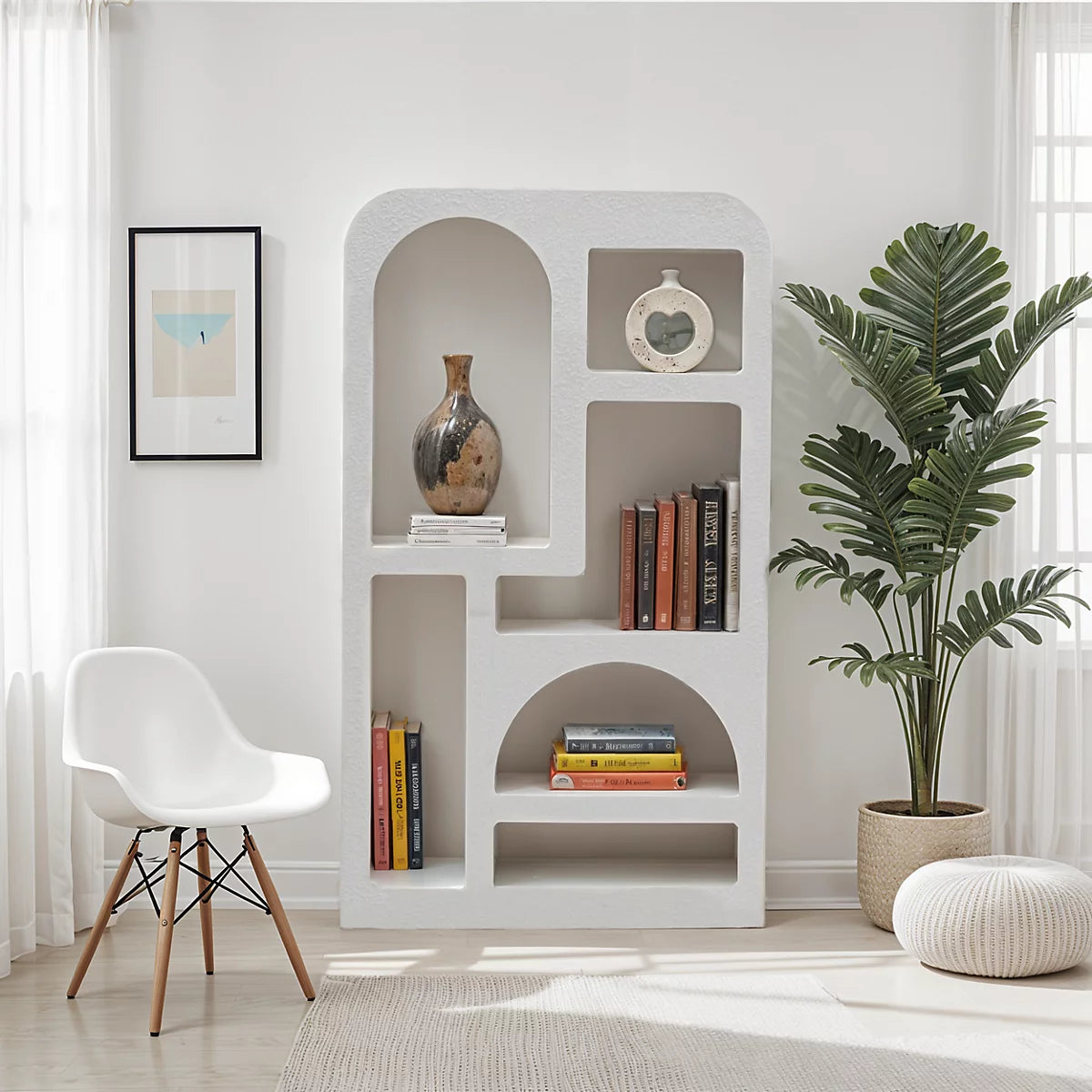 A modern living room with a Minimalist Cream Wind Sculptural Shelf displaying books, a vase, and a clock, complemented by a white chair, framed art, and a potted plant near the window.