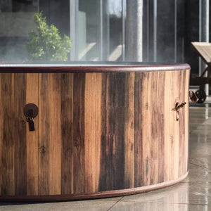 A close-up of the Outdoor Spa Hot Tub, a round cedar hot tub with steam rising from the water, positioned on a stone patio near glass doors to create the perfect outdoor spa experience.