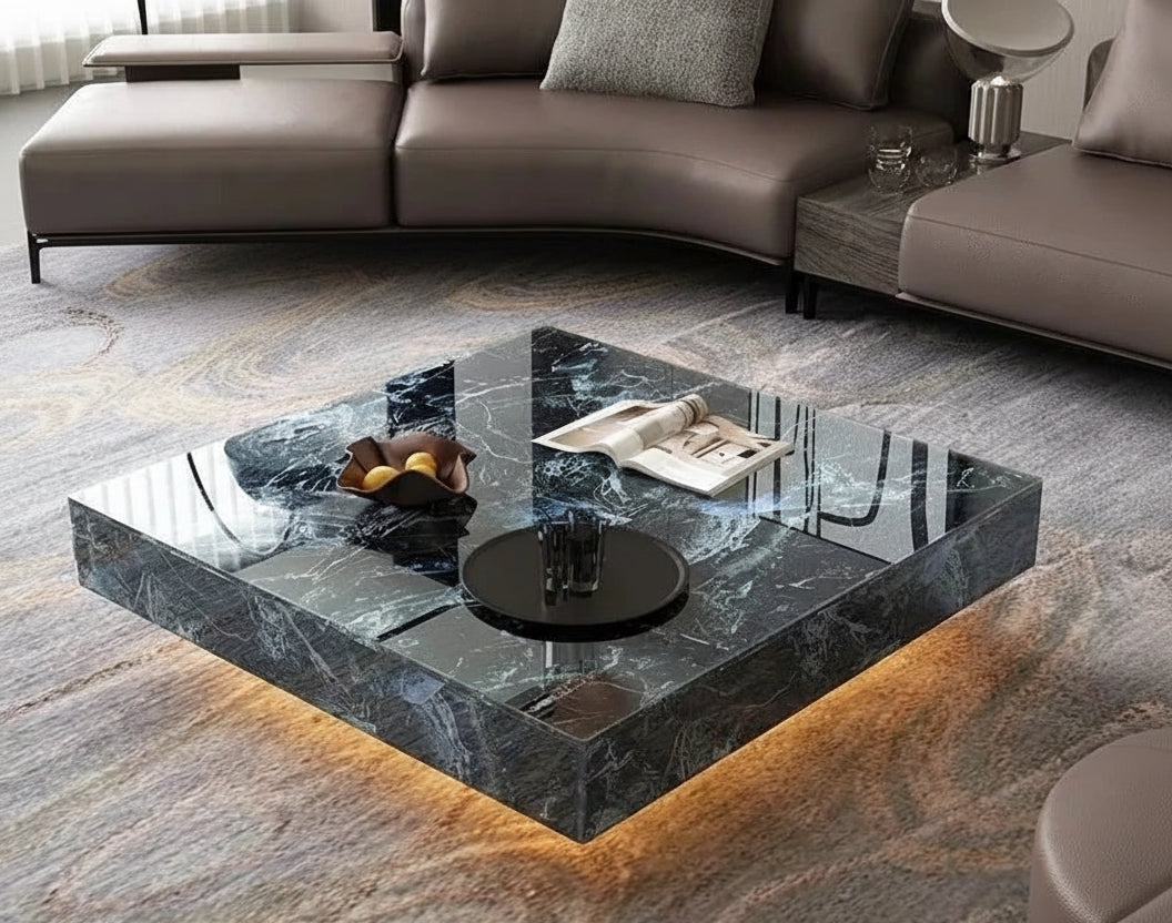 A modern living room features the Modern Luxe Coffee Table with a bowl of fruit, books, and glasses on it, set atop a patterned rug and complemented by a sectional sofa in the background.