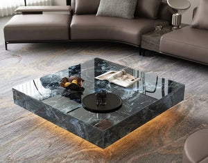 A modern living room features the Modern Luxe Coffee Table with a bowl of fruit, books, and glasses on it, set atop a patterned rug and complemented by a sectional sofa in the background.