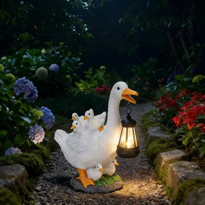 The Solar Duck Lamp stands on a stone garden path among blooming flowers at night, holding a solar-powered lantern and surrounded by gosling statues.