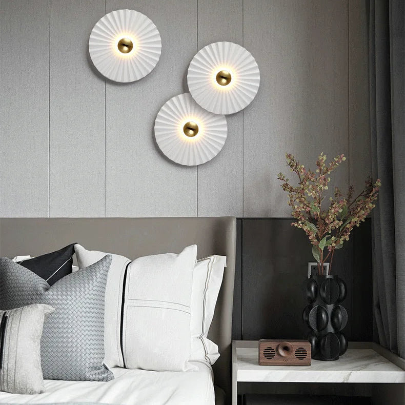 A modern bedroom with a grey accent wall features three Origami Circle Wall Lights above the bed, plus pillows and a nightstand adorned with a decorative vase and sculpture.