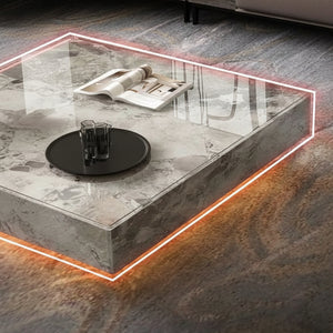 The Modern Luxe Coffee Table features a square marble base, underlit orange LED edges, a glass top with two glasses on a black tray, and a magazine—perfect for modern interiors and displayed on a textured carpet.
