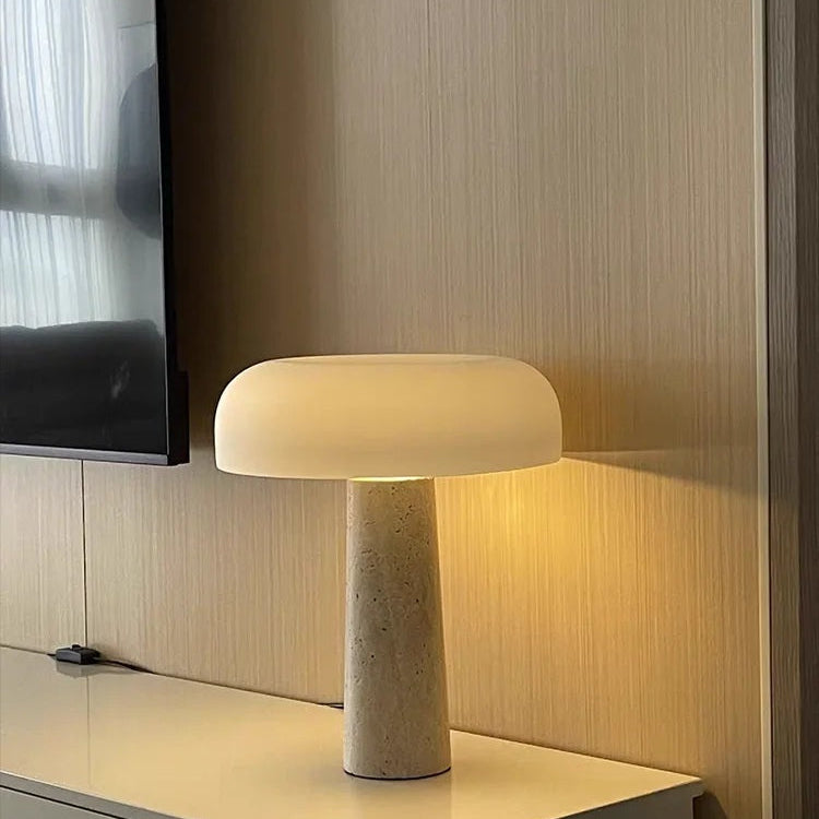 The Mushroom Desk Lamp, featuring a cylindrical stone base and a wide, rounded white shade, is illuminated and situated on a glossy surface beside a wall-mounted TV, providing organic lighting for any modern space.