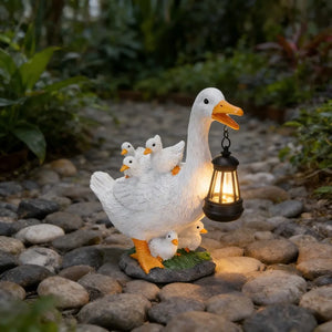 The Solar Duck Lamp features a charming duck figurine holding a solar-powered lantern, surrounded by ducklings and set on a stone garden path with lush greenery in the background.