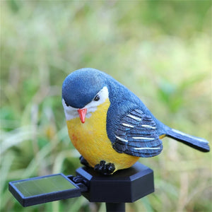 The Solar Bird Light features a realistic songbird decor figurine in blue, yellow, and white perched on a black stand with a small solar panel, making it the perfect garden solar light accent against a blurred green background.
