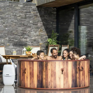 Four people relax together in the Outdoor Spa Hot Tub, placed outside near a modern house with dark stone walls and patio furniture in the background.
