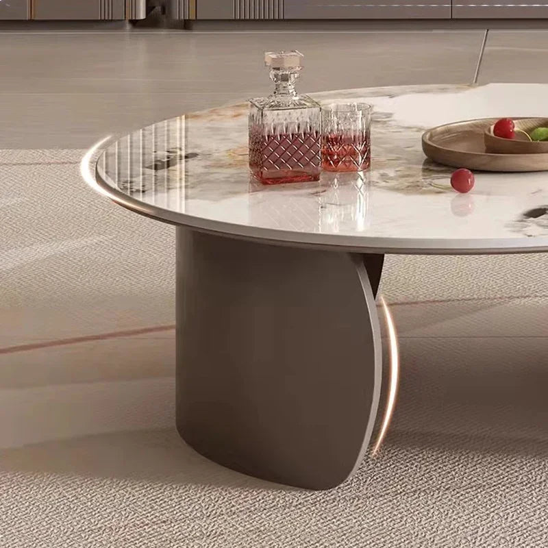 The Luxe Round Coffee Table with curved gray legs displays a crystal decanter, two matching glasses of dark liquid, and a wooden tray holding apples.