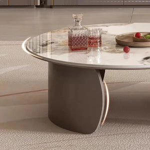 The Luxe Round Coffee Table with curved gray legs displays a crystal decanter, two matching glasses of dark liquid, and a wooden tray holding apples.