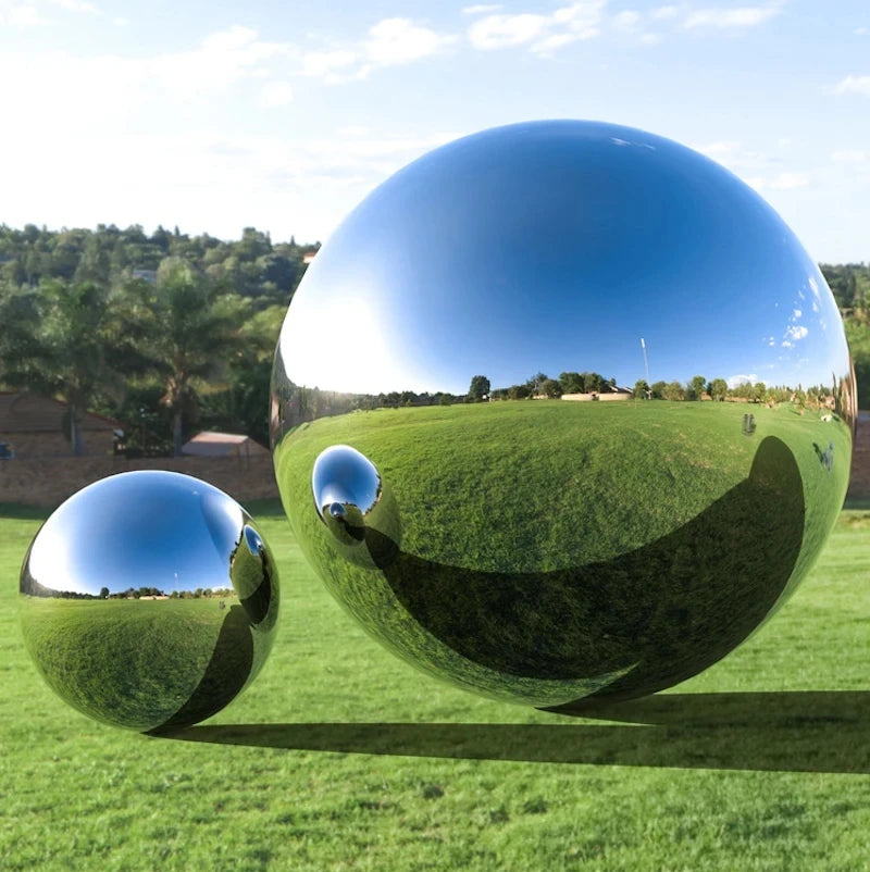 Two large Stainless Steel Gazing Balls with a reflective mirror polish rest on a grassy field, mirroring the sky, trees, and landscape as stunning garden decorations.