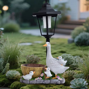 Adding whimsy to your garden, the Solar Goose Lamp features charming duck and duckling figurines, a small bowl, succulents, and flowers that evoke a delightful cottage garden feel, all set before a softly blurred house and steps in the background.