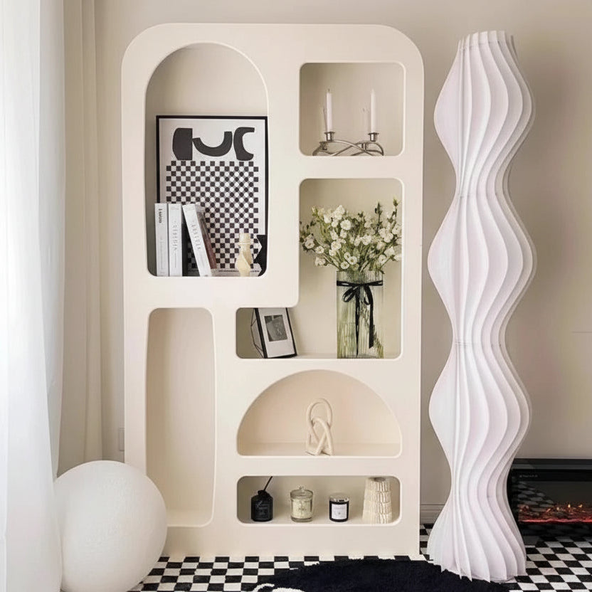 The Minimalist Cream Wind Sculptural Shelf, styled with books, decor, candles, and flowers, stands beside a tall wavy white floor lamp on a black-and-white checkered floor, capturing the French cream wind modern aesthetic.