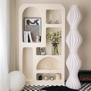 The Minimalist Cream Wind Sculptural Shelf, styled with books, decor, candles, and flowers, stands beside a tall wavy white floor lamp on a black-and-white checkered floor, capturing the French cream wind modern aesthetic.