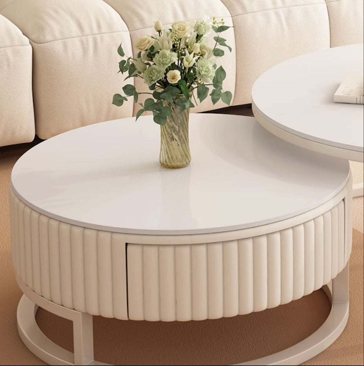 The Luxury Round Coffee Table Set, with its white minimalist design and ribbed base, holds a vase of white and light green flowers elegantly in front of a beige sofa.