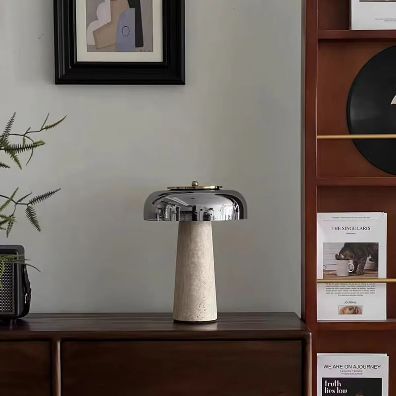 The Mushroom Desk Lamp, featuring a modern design with a metallic dome shade, is placed on a wooden console beside a plant, with shelves and framed art visible in the background.