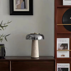 The Mushroom Desk Lamp, featuring a modern design with a metallic dome shade, is placed on a wooden console beside a plant, with shelves and framed art visible in the background.