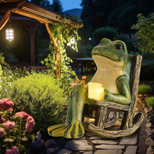 The Solar-powered Rocking Chair Frog Light features a large garden frog ornament statue sitting in a rocking chair and holding a glowing lantern amidst flowers and foliage at dusk. Crafted from weatherproof resin, this charming solar-powered light adds whimsical charm and gentle illumination to your outdoor space.