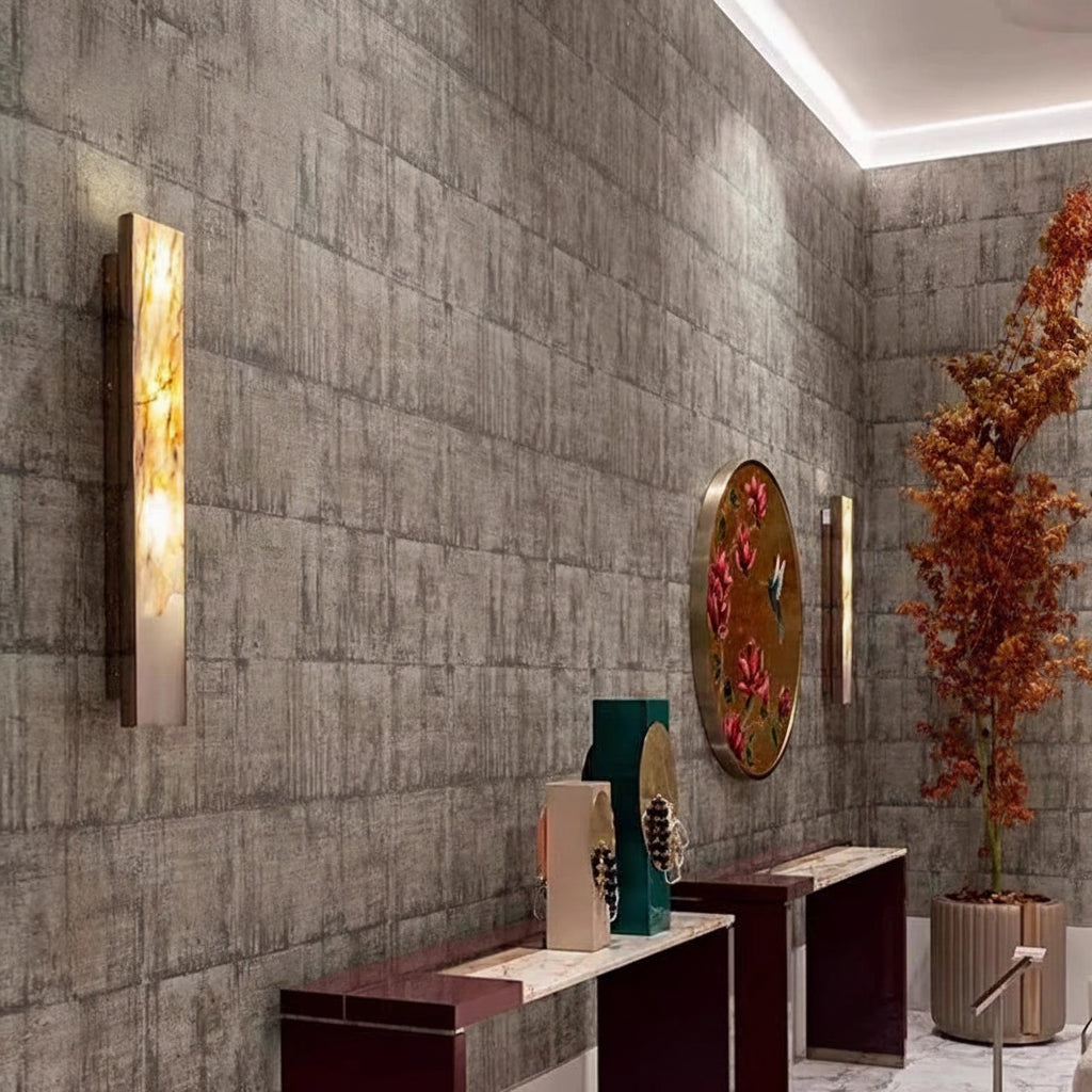 Modern hallway with textured gray walls, Luxury Marble Wall Sconces casting a warm glow, decorative vases on marble tables, a round wall art piece, and a tall plant with orange leaves in the corner.